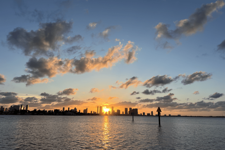 Sunset over a city skyline with clouds and a calm body of water in the foreground.