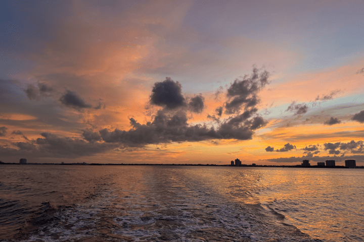 Sunset over water with silhouetted city skyline and clouded sky.