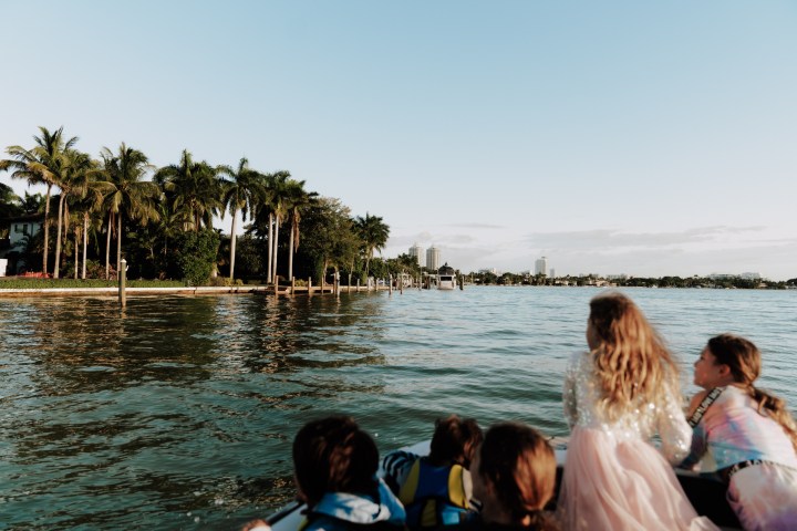 Children on a boat looking at palm trees and buildings along a waterfront.