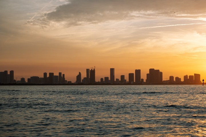 City skyline at sunset with water in foreground, orange sky and silhouetted buildings.